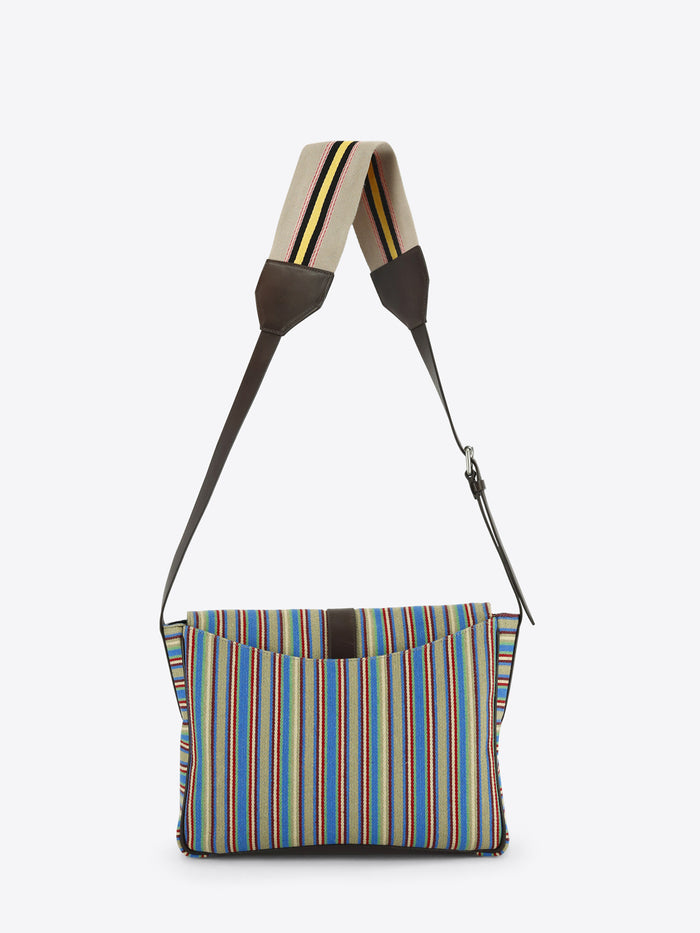 Back view of a multicolored striped shoulder bag with dark brown leather trim, a front buckle closure and an adjustable striped strap, shown against a light grey background.
