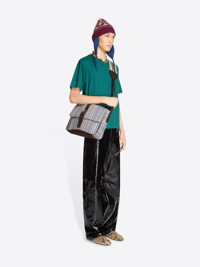 Model wearing a teal T-shirt, glossy black trousers, a burgundy patterned earflap beanie and snakeskin-print shoes, carrying a multicolored striped crossbody bag against a light grey background.