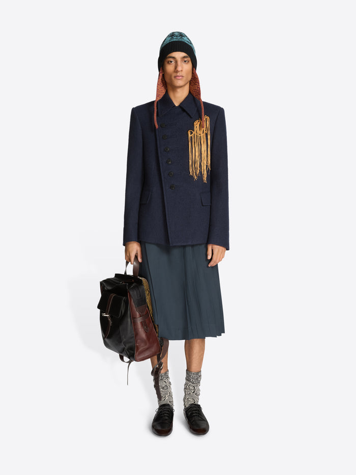 Person wearing a dark coat, gray skirt, and patterned shoes holding a backpack on a light gray background