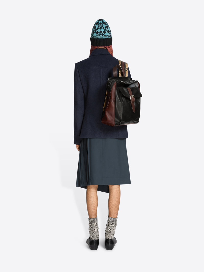 A model shown from behind wearing a navy coat, dark gray skirt, patterned socks, and a blue-and-black patterned beanie, carrying a brown and black leather shoulder bag against a light gray background.