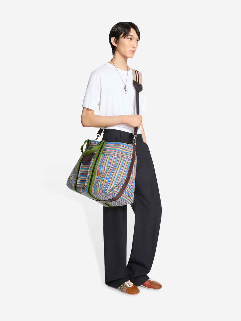 A model wearing a white t-shirt, black wide-leg trousers, and brown loafers, carrying a striped shoulder bag a light gray background.