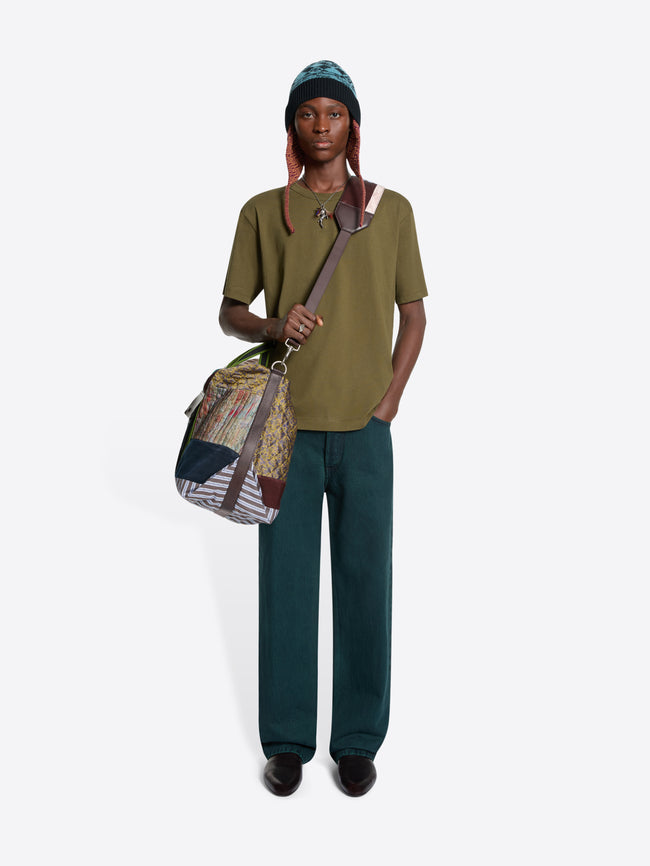 A model wearing an olive green t-shirt, dark teal trousers, a green beanie, and holding a large, colorful patchwork shoulder bag with green and striped details, set against a light gray background.