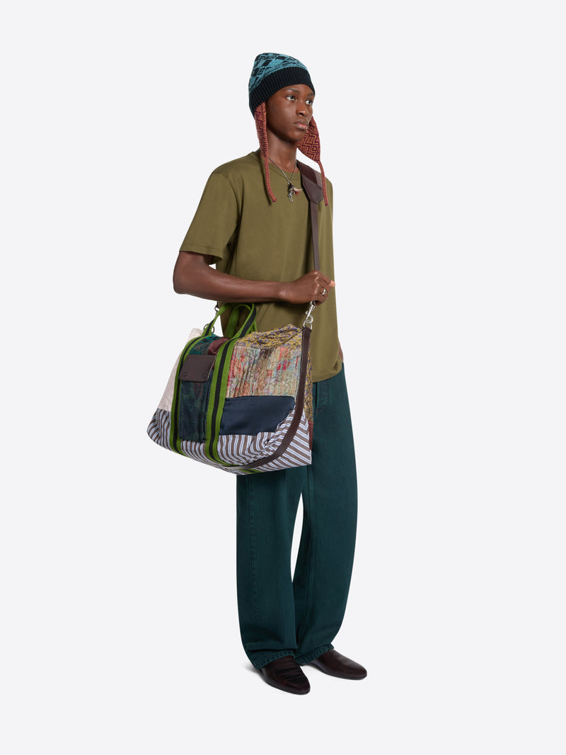 A model wearing an olive green t-shirt, dark teal trousers, a green beanie, and holding a large, colorful patchwork shoulder bag with green and striped details, set against a light gray background.