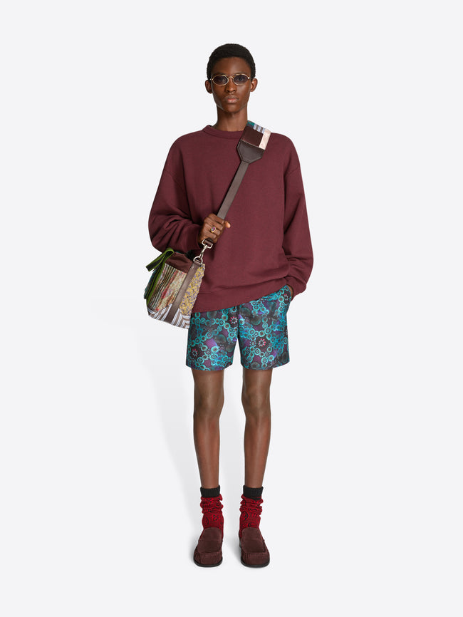 Person wearing a burgundy sweatshirt, patterned shorts, and brown shoes on a white background, holding a patchwork tote bag.