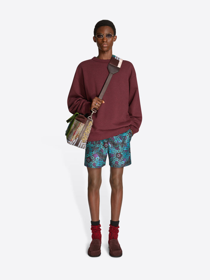 Person wearing a burgundy sweatshirt, patterned shorts, and brown shoes on a white background, holding a patchwork tote bag.