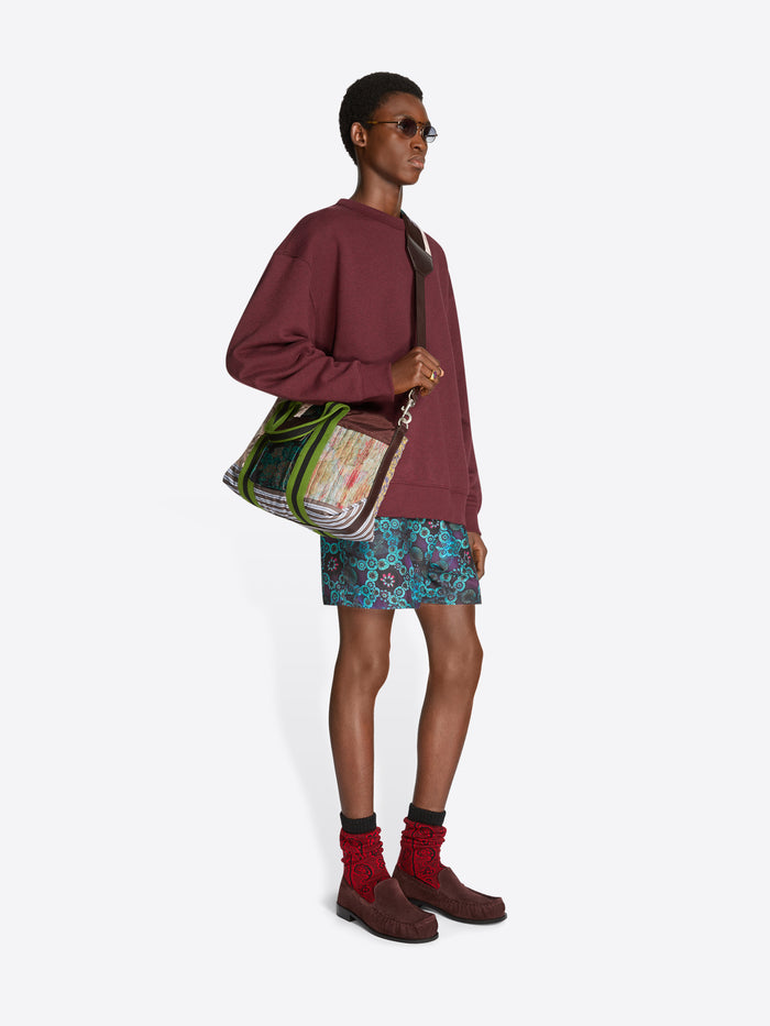 Person wearing a burgundy sweatshirt, patterned shorts, and brown shoes on a white background, holding a patchwork tote bag.