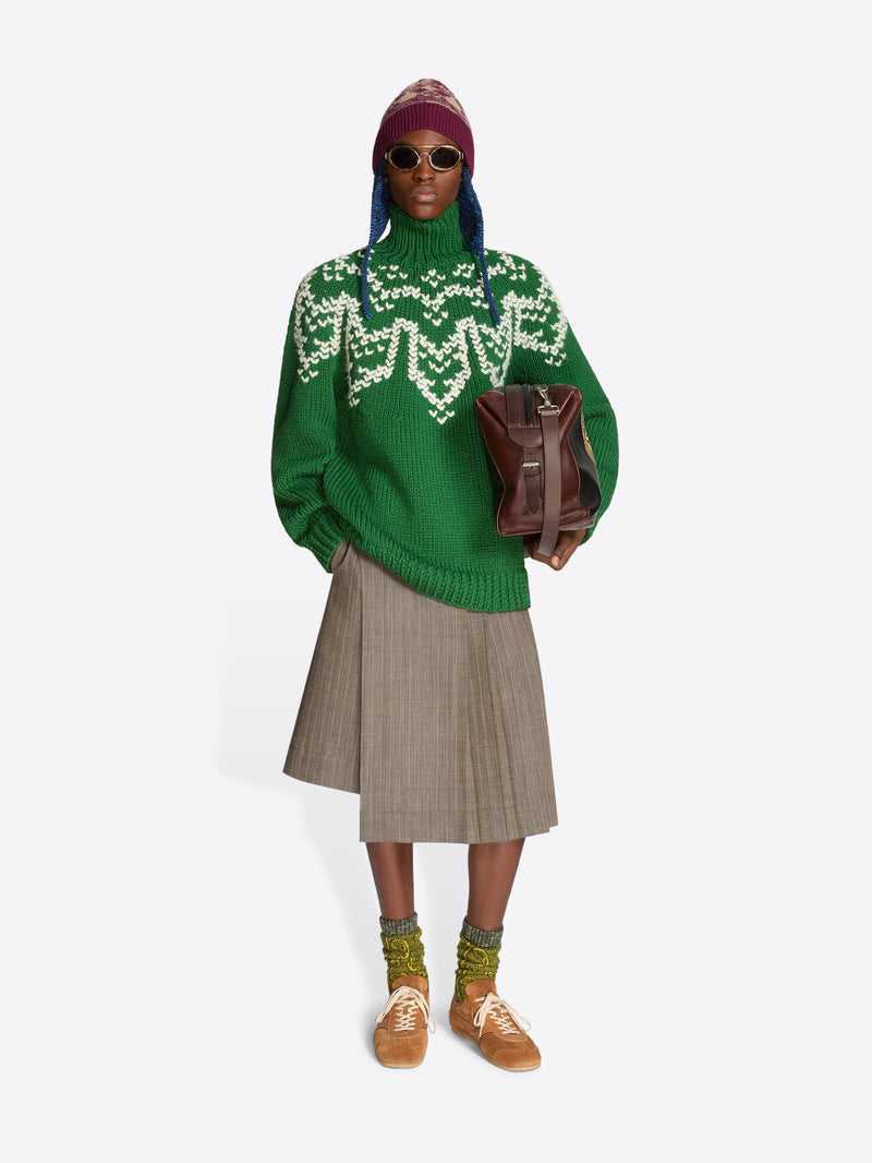 A model stands against a light gray background wearing a green knit turtleneck sweater with a white geometric pattern across the chest, a pleated taupe skirt, brown suede sneakers, a burgundy hat, and holding a brown leather bag.