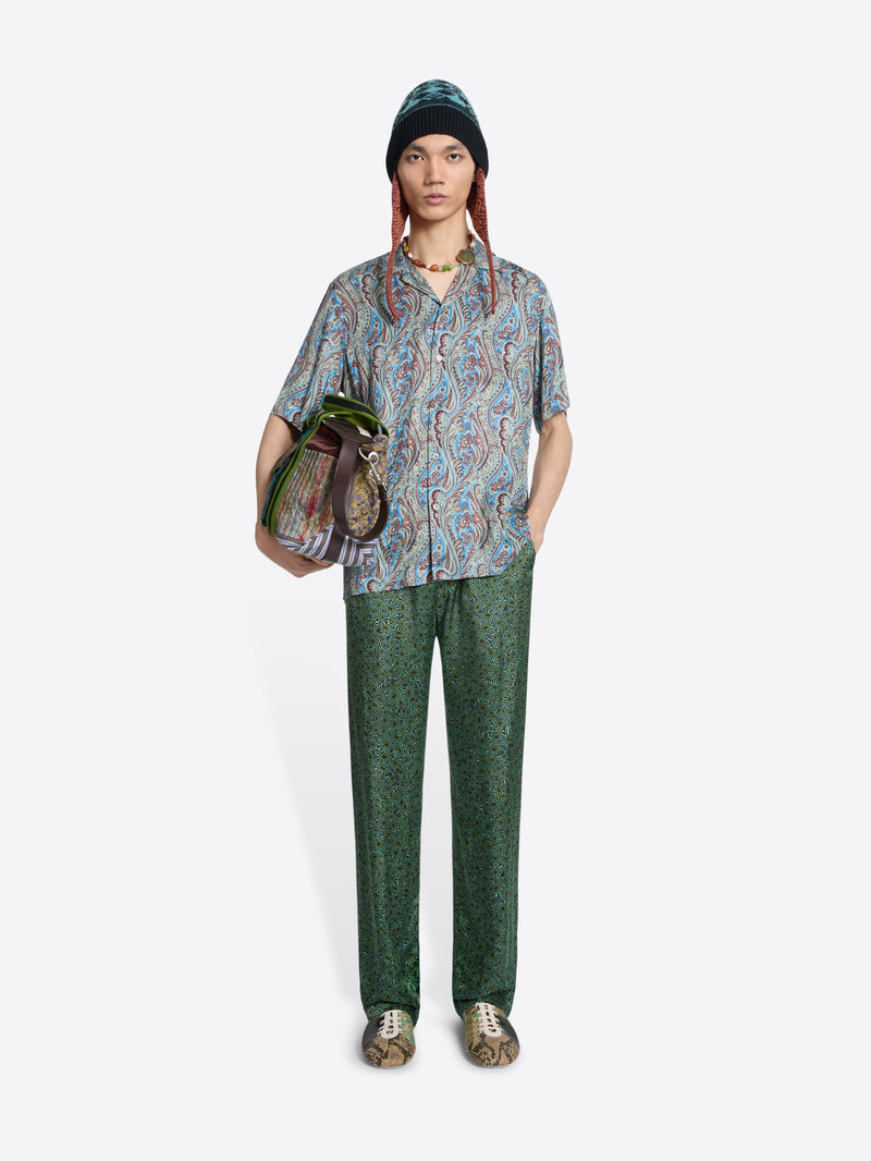 Person wearing a patterned shirt and green pants with a colorful bag on a light gray  background