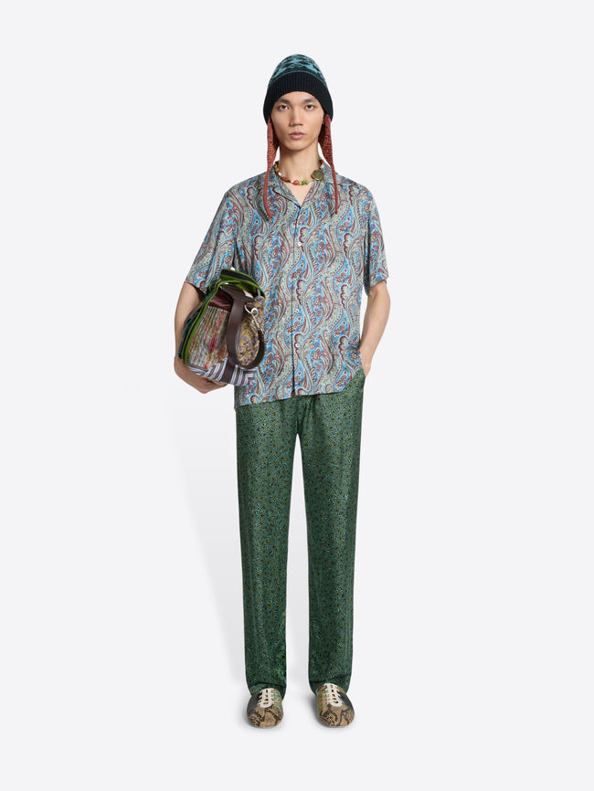 Person wearing a patterned shirt and green pants with a colorful bag on a light gray  background