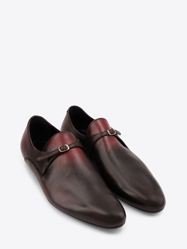 A pair of dark brown leather shoes with a single strap and a small silver buckle across the top, displayed on a white background.