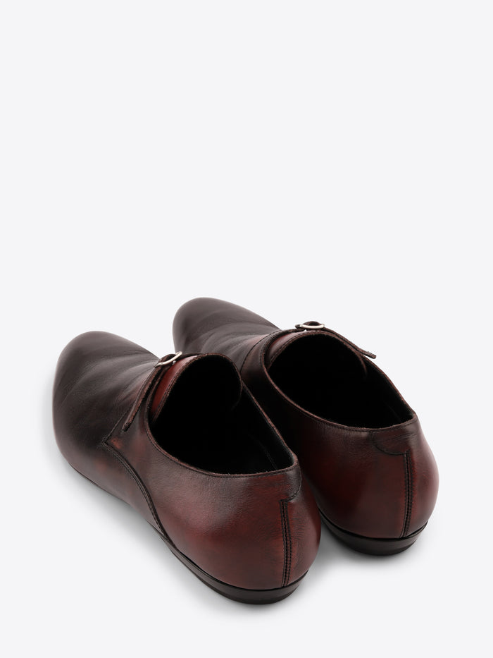 A pair of dark brown leather shoes with a single strap and a small silver buckle across the top, displayed on a white background.