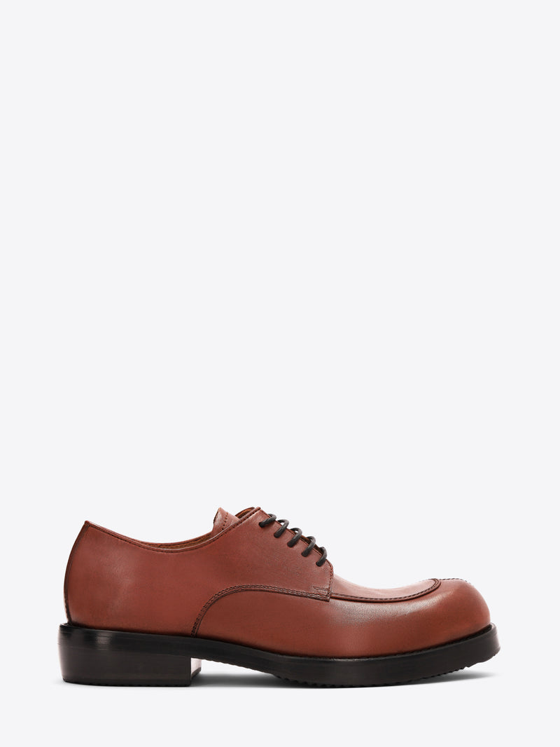 Reddish-brown leather lace-up shoe with an apron toe, chunky black sole, and low heel on a light gray background.