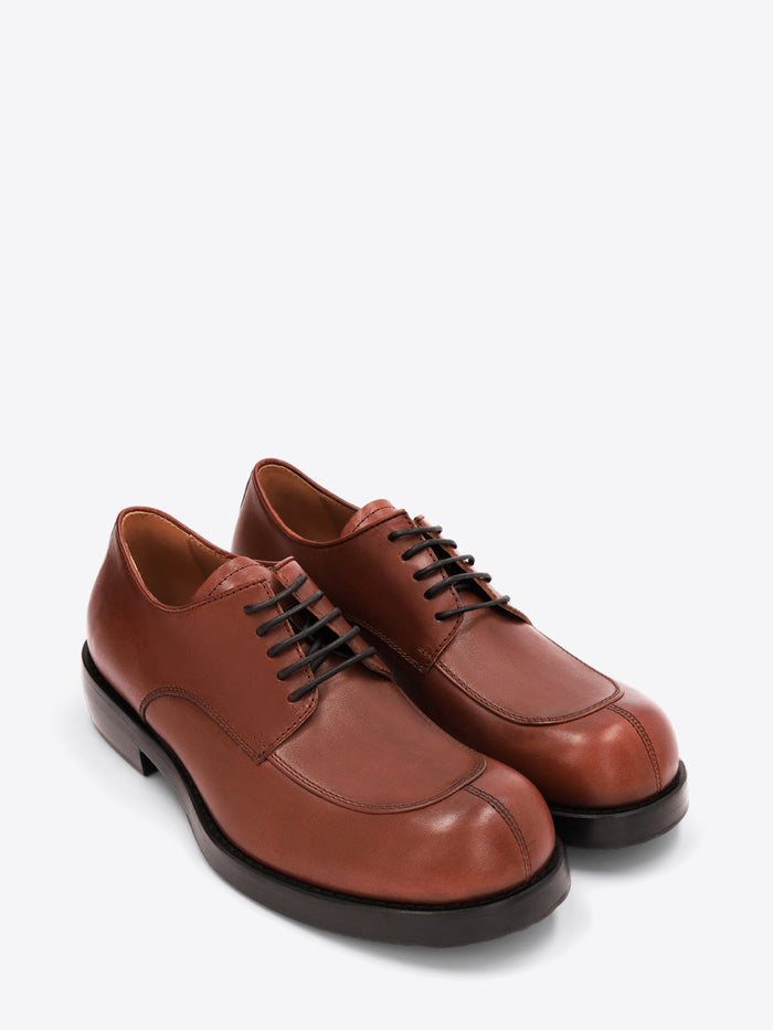 A pair of men's reddish-brown leather lace-up shoes with an apron toe, chunky black sole, and low heel on a light gray background.