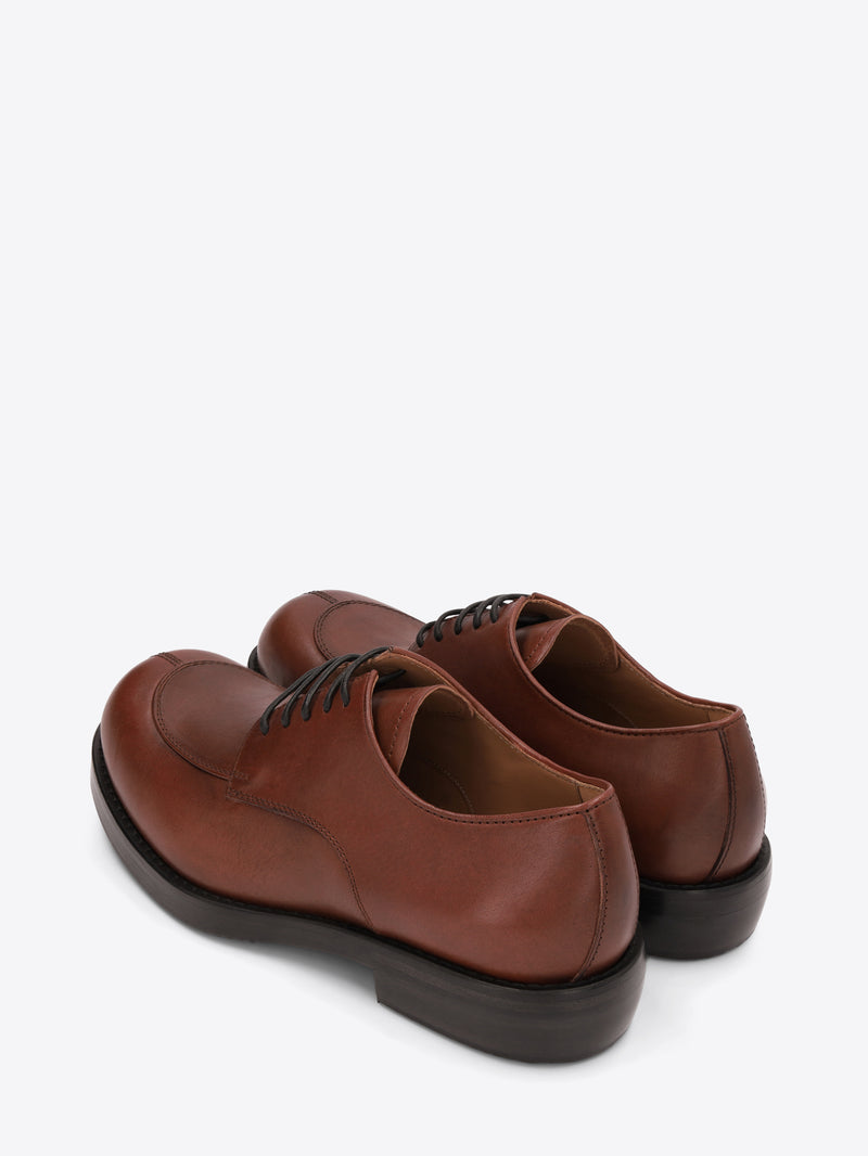 A pair of men's reddish-brown leather lace-up shoes with an apron toe, chunky black sole, and low heel on a light gray background.