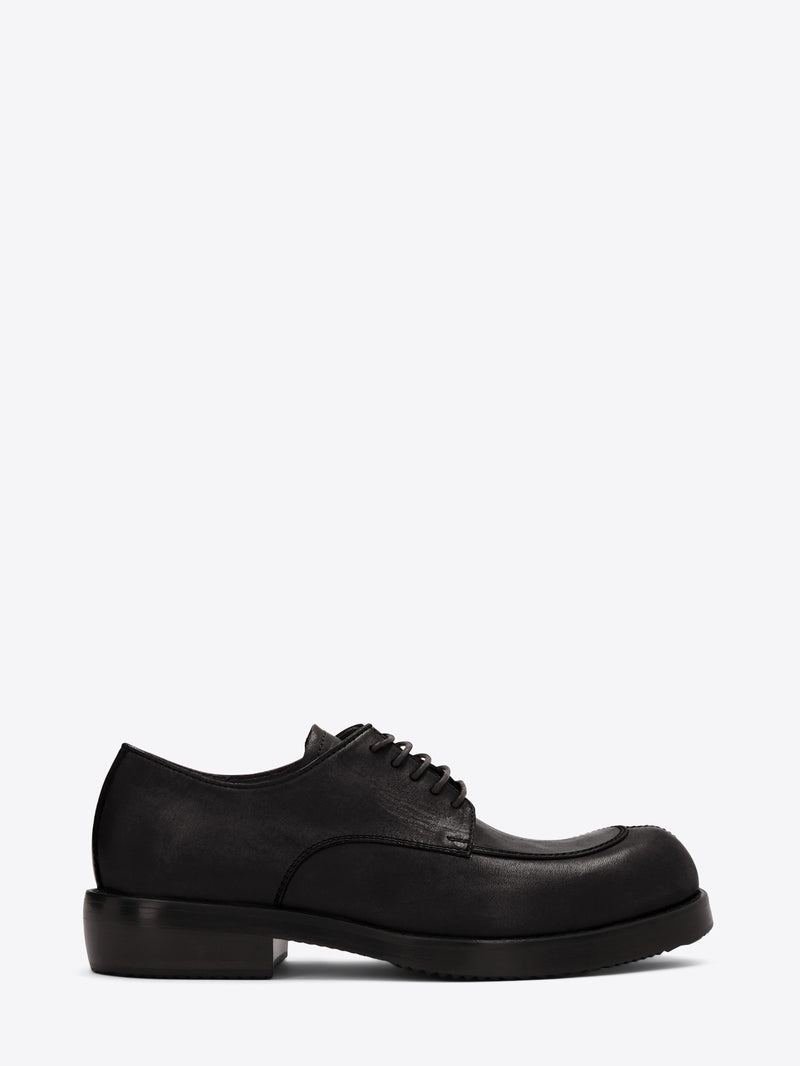 Black leather lace-up shoe with an apron toe, chunky sole, and low stacked heel on a light gray background.