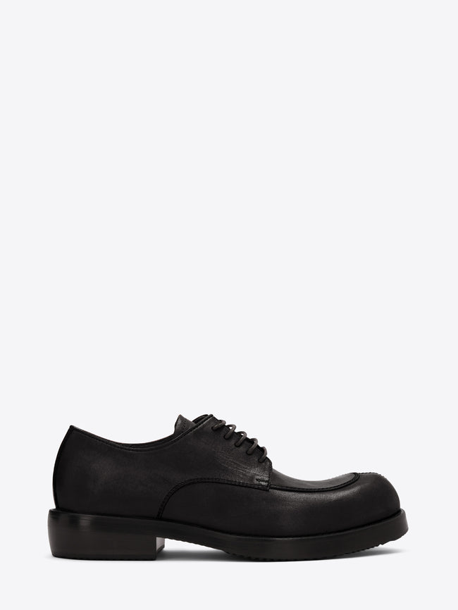 Black leather lace-up shoe with an apron toe, chunky sole, and low stacked heel on a light gray background.