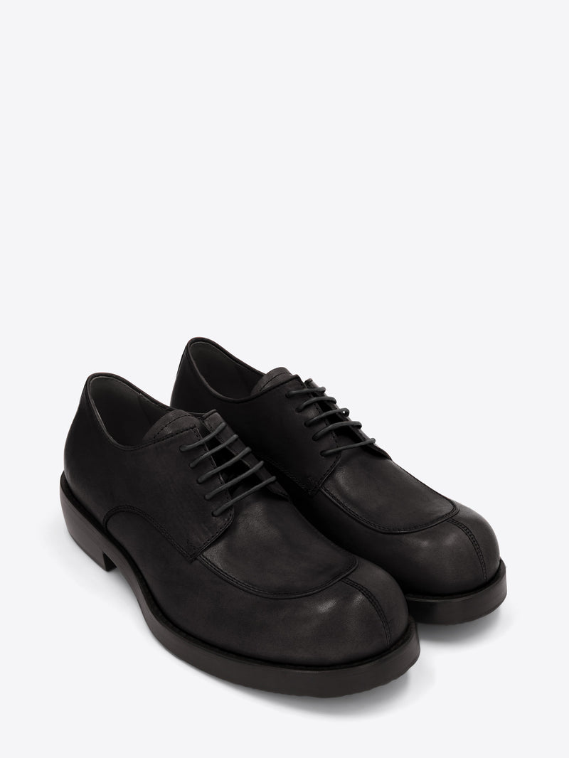 A pair of black leather lace-up shoes with an apron toe, chunky sole, and low stacked heel on a light gray background.