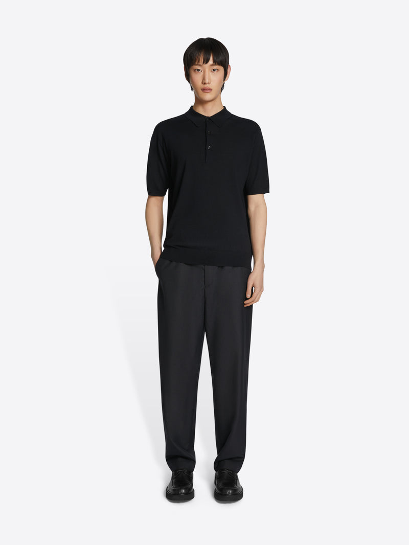 A model stands against a light gray background wearing a black short-sleeve polo shirt with a partial button placket and black wide-leg trousers.