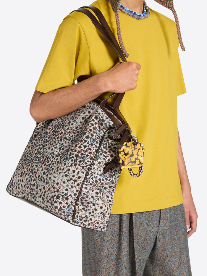 A person wearing a bright yellow t-shirt and gray trousers carries a large floral-print shoulder bag with brown leather straps and a matching small yellow pouch key chain. 