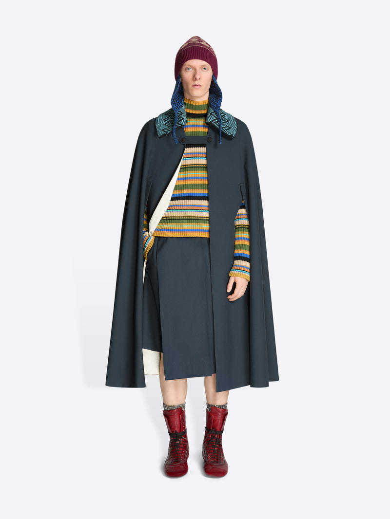 Person wearing a dark gray cape, knitted detachable collar, colorful striped sweater, and red boots on a white background
