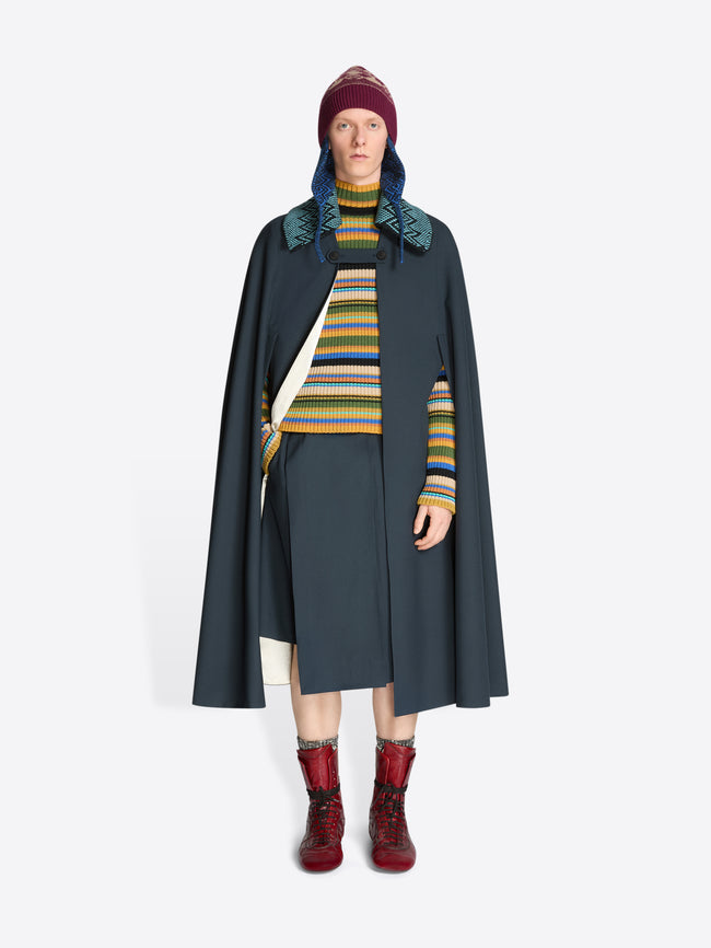 Person wearing a dark gray cape, knitted detachable collar, colorful striped sweater, and red boots on a white background