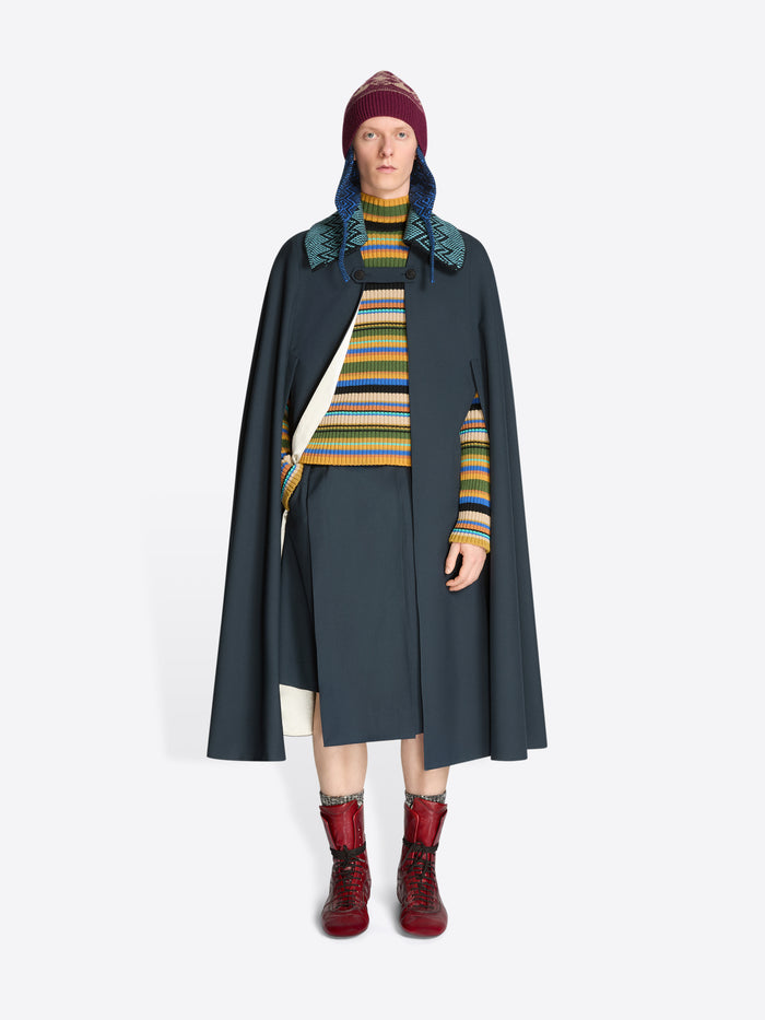 Person wearing a dark gray cape, knitted detachable collar, colorful striped sweater, and red boots on a white background