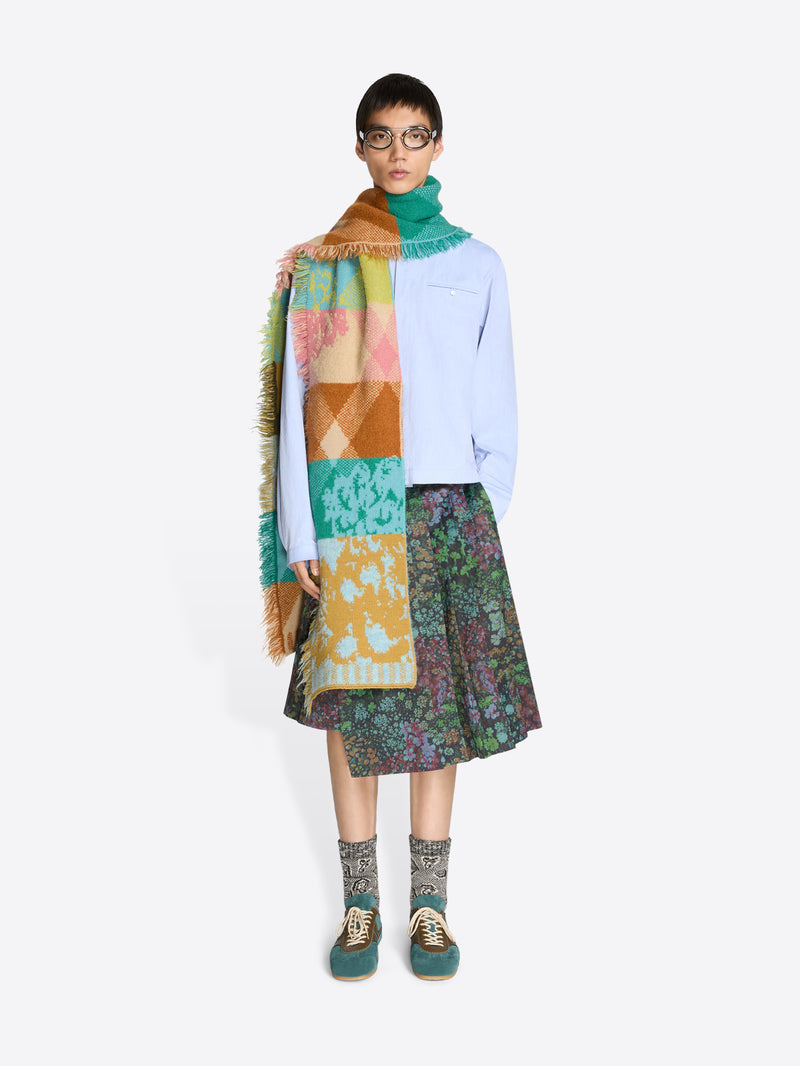 Person wearing a colorful scarf, blue shirt, patterned skirt, and patterned socks on a white background
