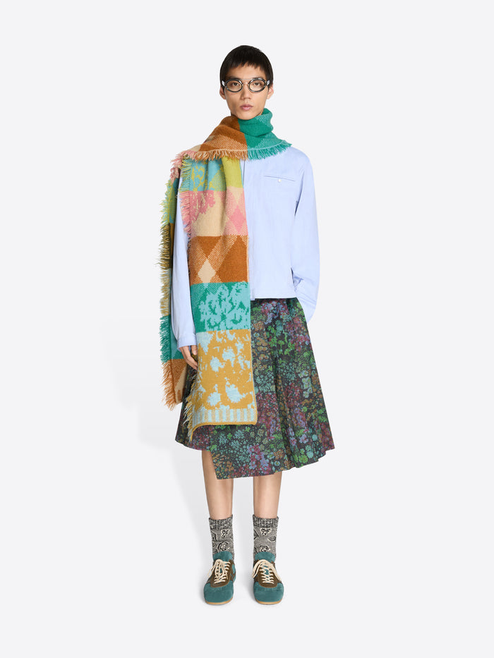 Person wearing a colorful scarf, blue shirt, patterned skirt, and patterned socks on a white background