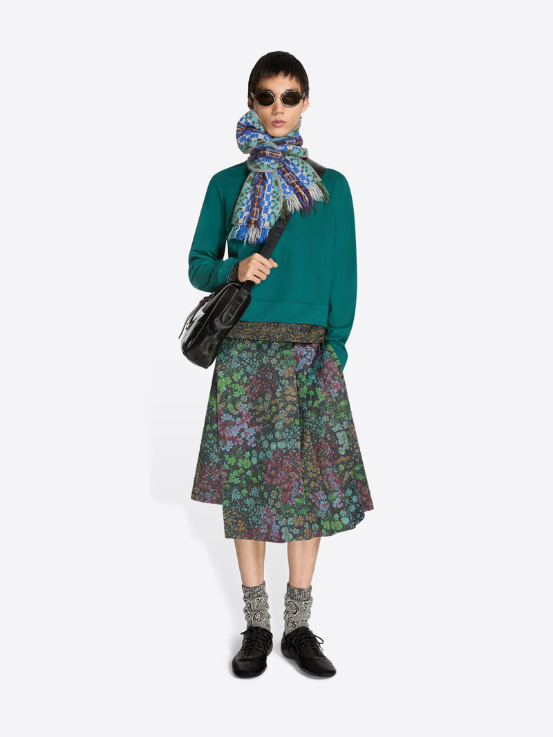 Person wearing a teal sweatshirt, patterned floral skirt, colorful scarf and sunglasses on a white background
