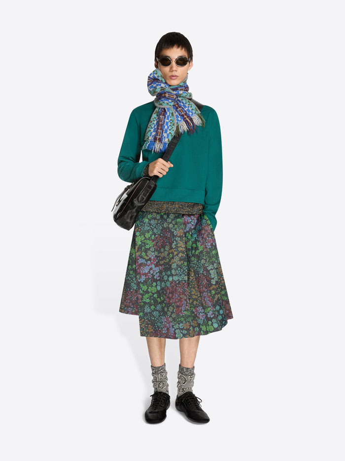 Person wearing a teal sweatshirt, patterned floral skirt, colorful scarf and sunglasses on a white background