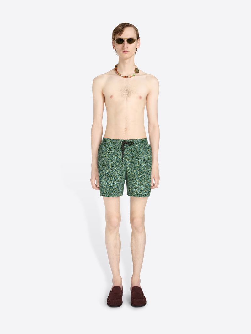 A male model stands against a light gray background wearing green patterned swim trunks, sunglasses, a beaded necklace, and dark loafers.