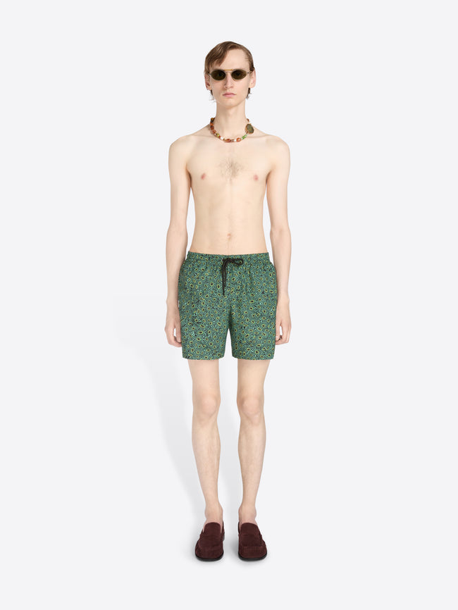 A male model stands against a light gray background wearing green patterned swim trunks, sunglasses, a beaded necklace, and dark loafers.