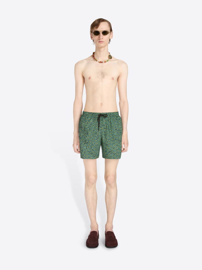 A male model stands against a light gray background wearing green patterned swim trunks, sunglasses, a beaded necklace, and dark loafers.