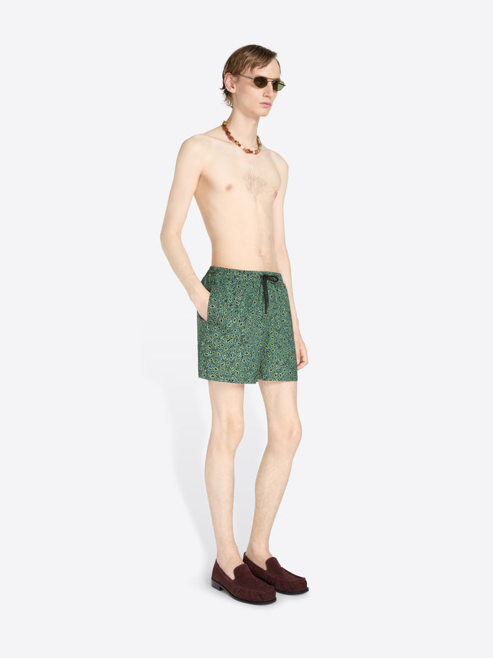 A male model stands against a light gray background wearing green patterned swim trunks, sunglasses, a necklace, and dark loafers.
