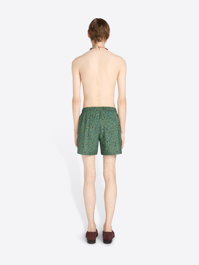 Back view of a male model standing against a light gray background wearing green patterned swim trunks, sunglasses, a necklace, and dark loafers.
