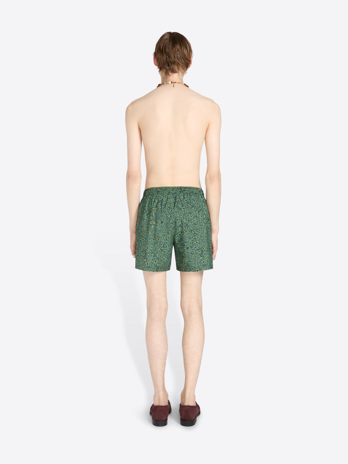 Back view of a male model standing against a light gray background wearing green patterned swim trunks, sunglasses, a necklace, and dark loafers.
