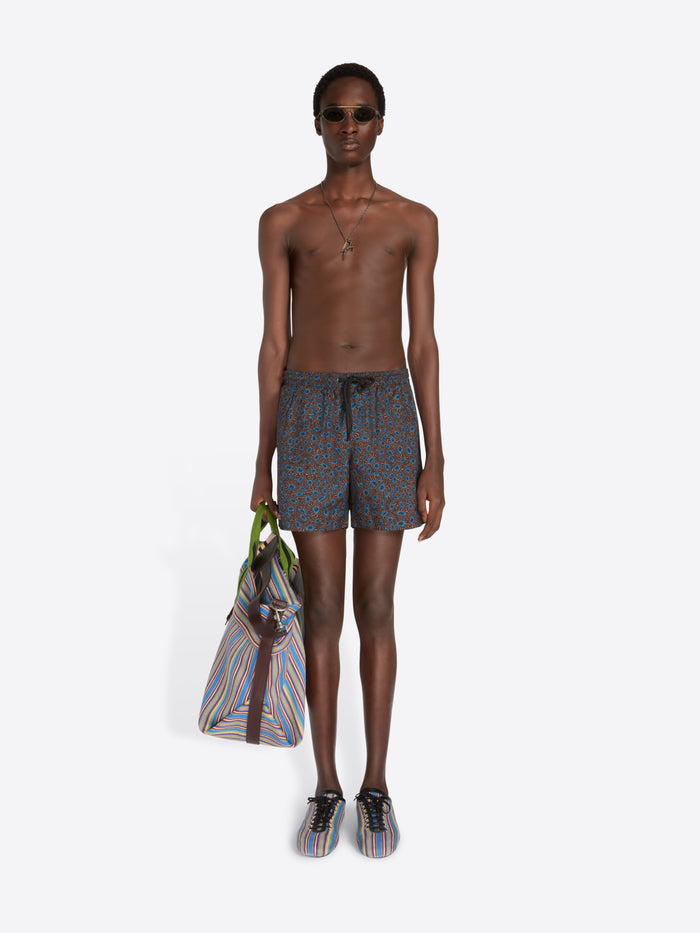 A male model stands against a light gray studio background wearing patterned blue-gray swim shorts, sunglasses, a necklace, and striped sneakers, while holding a striped tote bag.