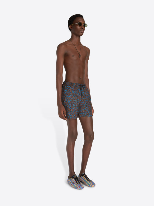 A male model stands against a light gray studio background wearing patterned blue-gray swim shorts, sunglasses, a necklace, and striped sneakers, while holding a striped tote bag.