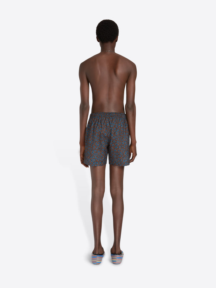 Back view of a male model standing against a light gray studio background wearing patterned blue-gray swim shorts, sunglasses, a necklace, and striped sneakers, while holding a striped tote bag.