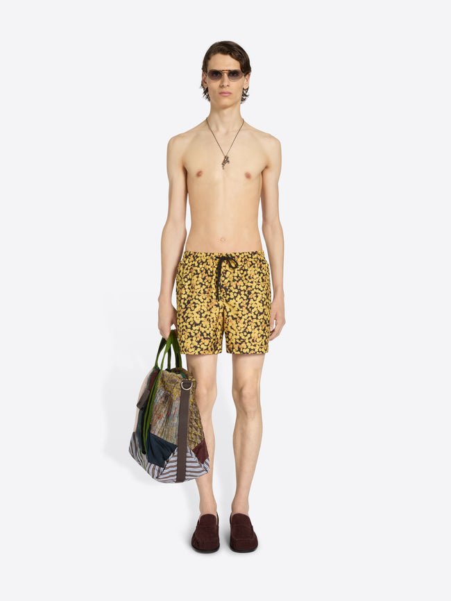 A pair of men's swim trunks featuring a dense yellow and black abstract floral, with a black drawstring at the elastic waistband, shown flat on a light gray background.