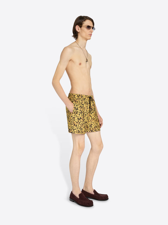 A male model wearing yellow and black swim trunks, styled with dark brown suede loafers and sunglasses, standing against a plain light gray background.
