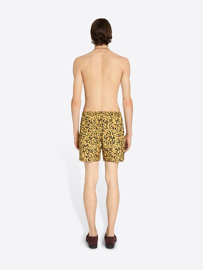 A man shown from the back wearing yellow and black patterned swim trunks, standing against a plain light gray background.