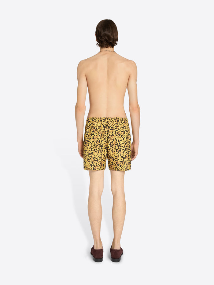 A man shown from the back wearing yellow and black patterned swim trunks, standing against a plain light gray background.