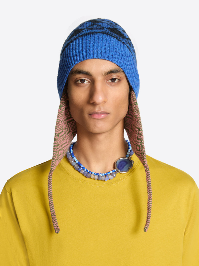 A model wearing a blue knit beanie with brown patterned ear flaps and a chunky blue beaded necklace, set against a plain light gray background.