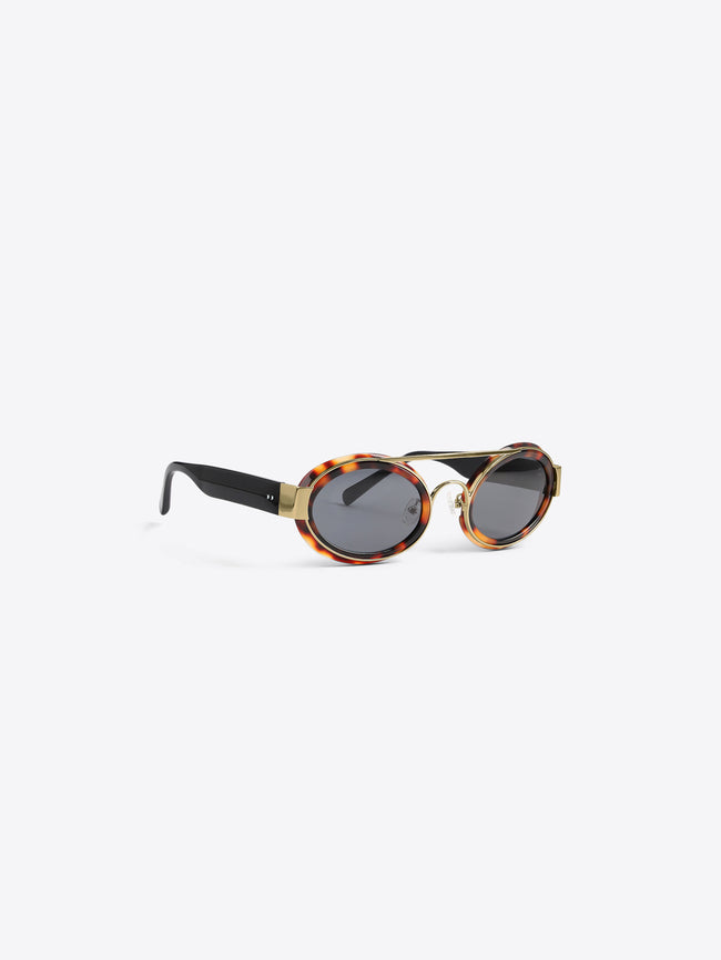A pair of round sunglasses with black temples, gold metal hardware, and a tortoiseshell-patterned acetate frame, shown from a side profile on a light gray background.