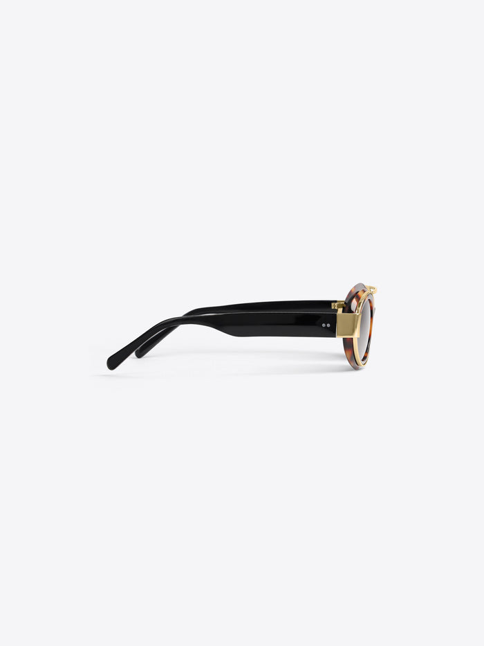 A pair of round sunglasses with black temples, gold metal hardware, and a tortoiseshell-patterned acetate frame, shown from a side profile on a light gray background.