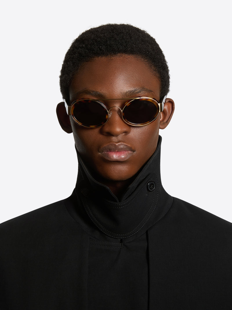 A close-up portrait of a model wearing oval sunglasses with gold frames and dark lenses, paired with a black high-neck top, against a light gray background.