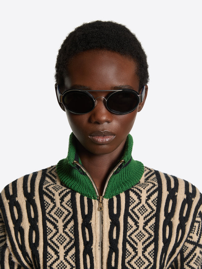 A model wearing black oval sunglasses and a beige and black patterned zip-up sweater with a green collar, against a light gray background.
