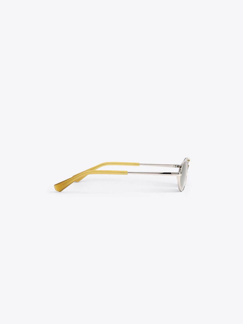 A pair of oval-frame sunglasses with translucent yellow acetate arms and silver metal temples, shown from a side profile against a light gray background. 