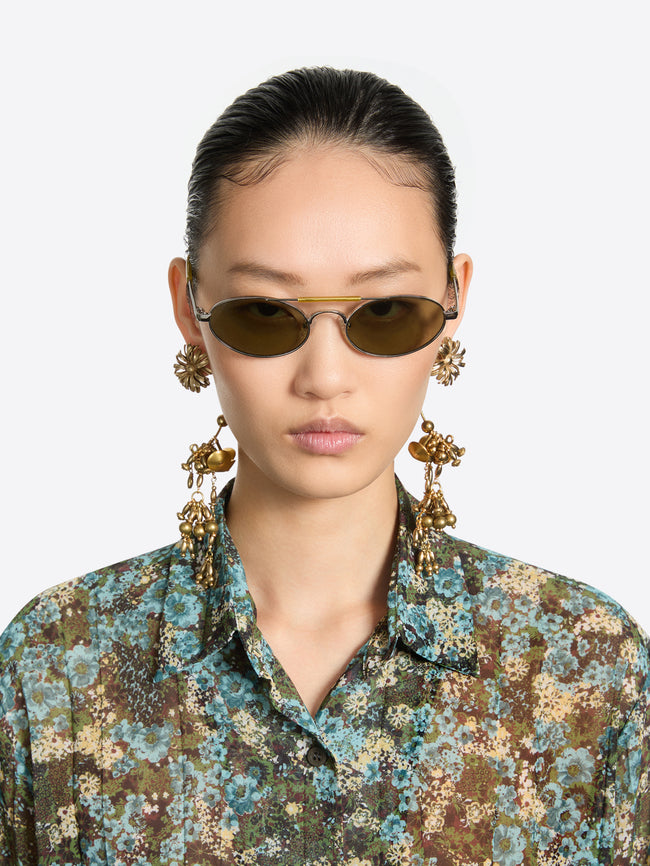 A model wearing oval-framed sunglasses with dark brown lenses, matching dangling earrings, and a blue and green floral patterned shirt, set against a light gray background.